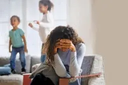 A mother holds her head in her hands as her children jump on the couch behind her
