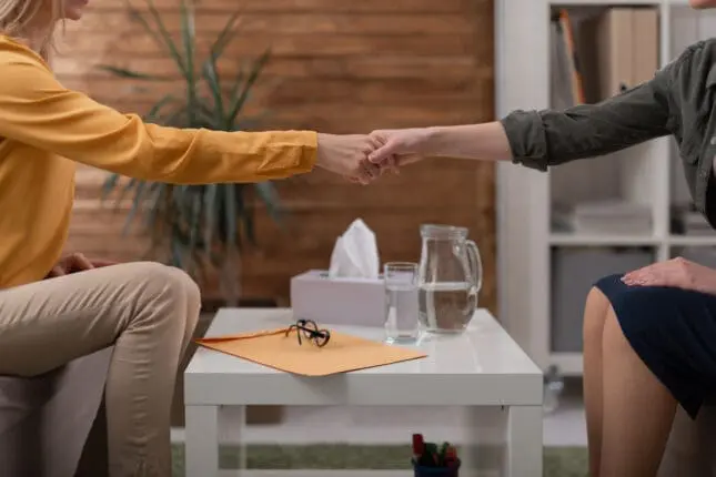 A therapist and client shake hands over a coffee table in the middle of the therapy room