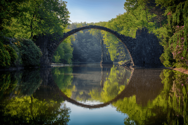 A bridge joining forests over a river, reflected in water.