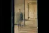 A blurry figure is visible through a doorway in an empty hallway