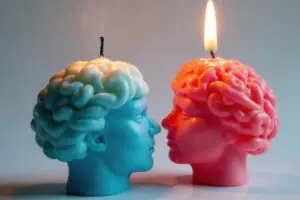 Two candles in the shape of human heads face each other; one is lit while the other is extinguished