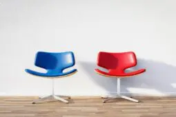Two empty seats, one red and one blue, sit beside each other against a blank wall