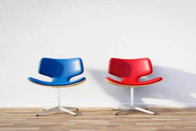 Two empty seats, one red and one blue, sit beside each other against a blank wall