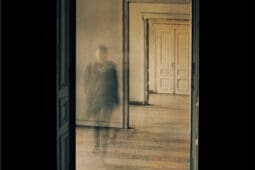 A blurry figure is visible through a doorway in an empty hallway
